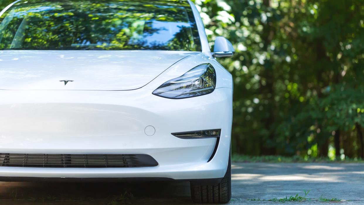   An all electric Tesla Model 3 in white on cement road with trees in background on sunny day. 