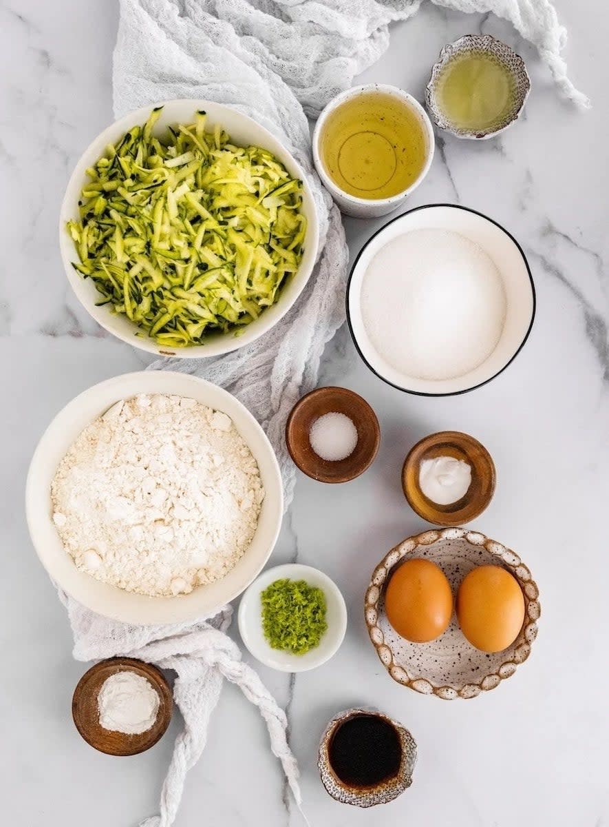 Ingredients on a marble countertop including shredded zucchini, flour, sugar, eggs, lemon zest, baking powder, salt, vanilla extract, and oil