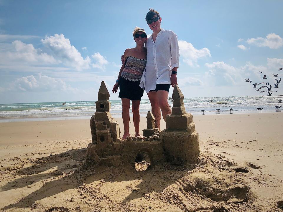 Two people posing behind a sandcastle on a beach.