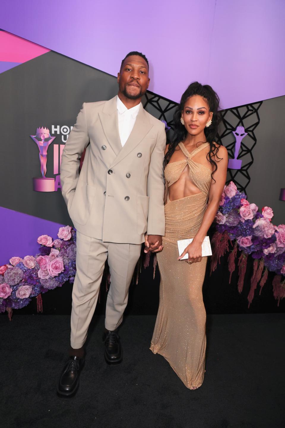 Jonathan Majors and Meagan Good at an event, Jonathan in a beige double-breasted suit, and Meagan in a tan cut-out gown, standing in front of a floral backdrop