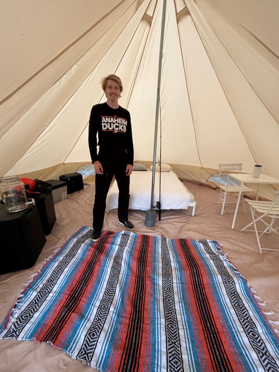 Oliver Russell stands in the middle of a large tent fitted with a table and chairs, rug, bed, and various other supplies.