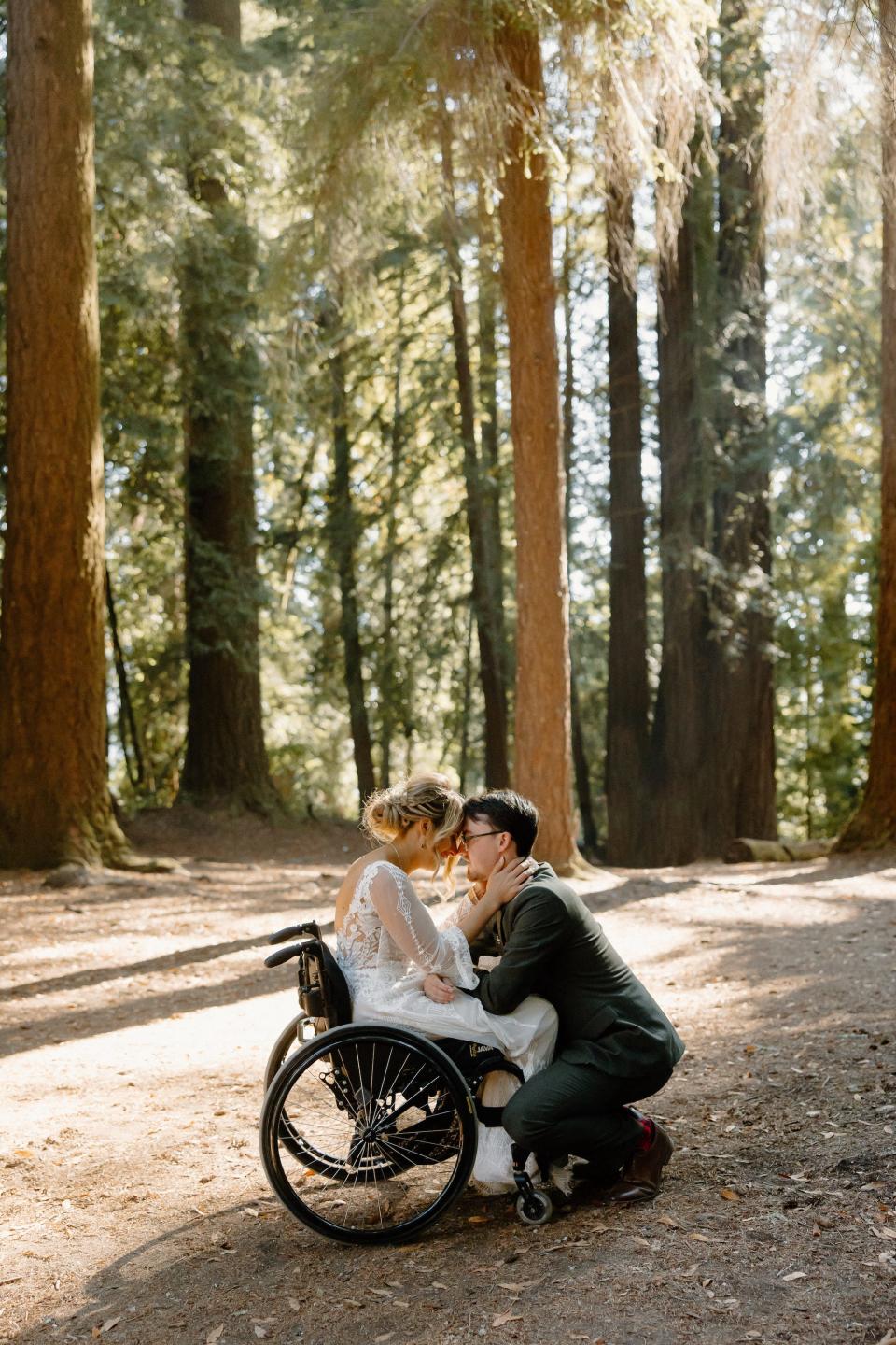 A groom kneels down to kiss his bride who sits in a wheelchair in a forest.