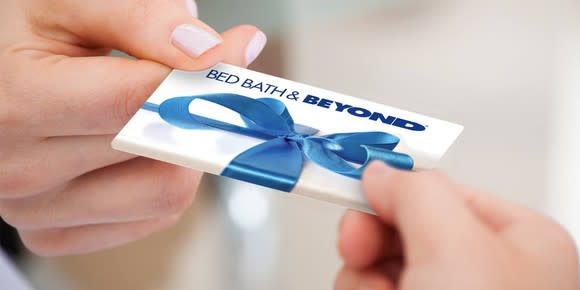 A hand giving a blue-and-white Bed Bath & Beyond gift card to another hand