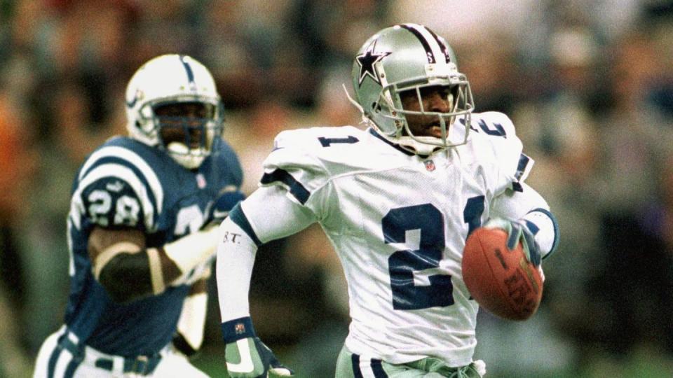 deion sanders runs while holding a football in his left hand, behind him is an opponent running after him, both men wear football uniforms and helmets