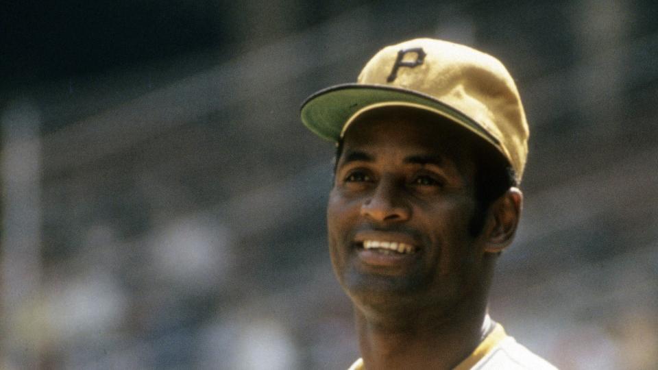 roberto clemente smiles and looks off into the distance, he wears a pittsburgh pirates uniform and yellow baseball cap and holds a baseball to his chest