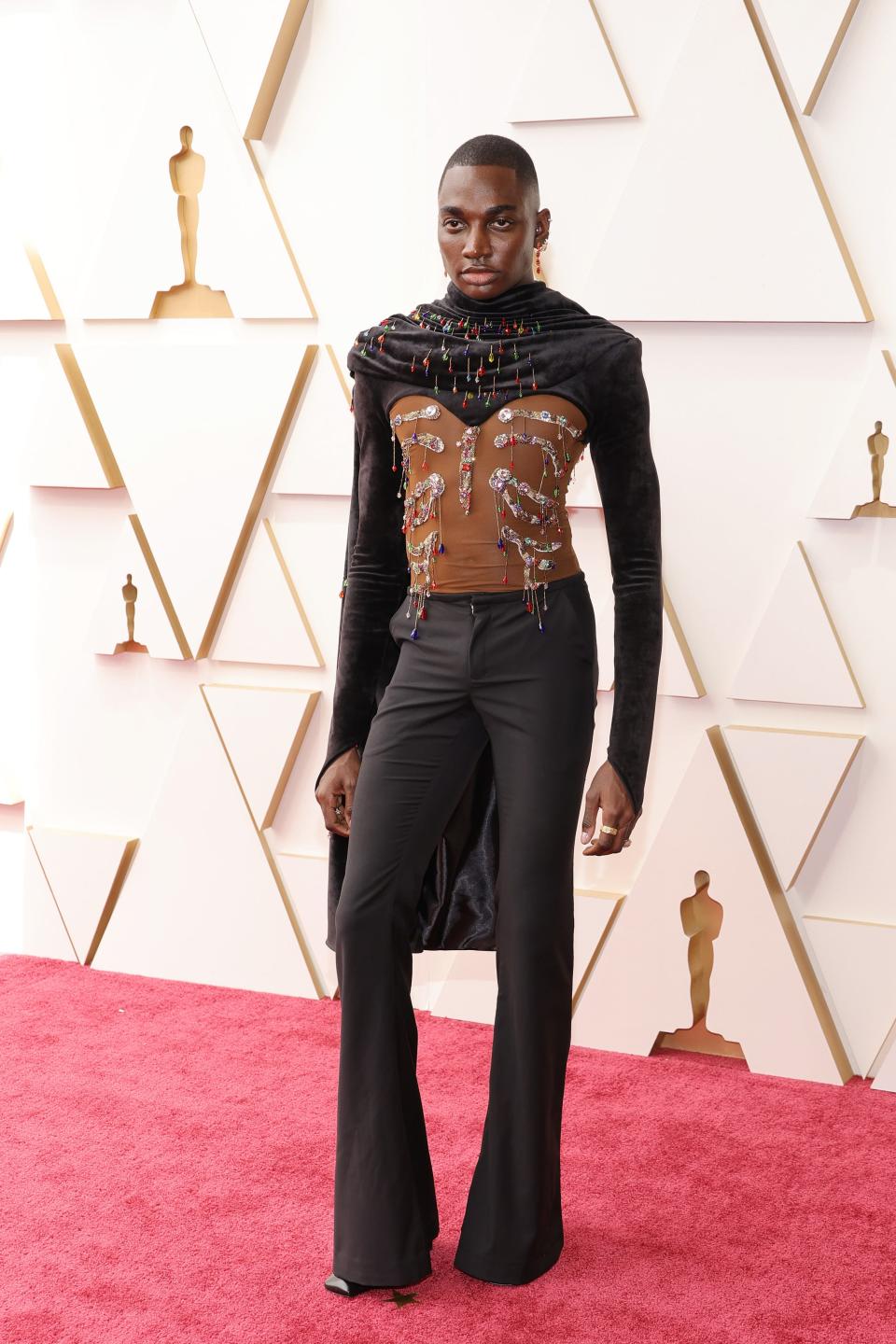 Rickey in a sheer bodice top with embellishments assembled to look like a rib cage. He's top his black long sleeves and a black cape with colorful crystals. He's wearing black flared trousers and square-toed black heels.