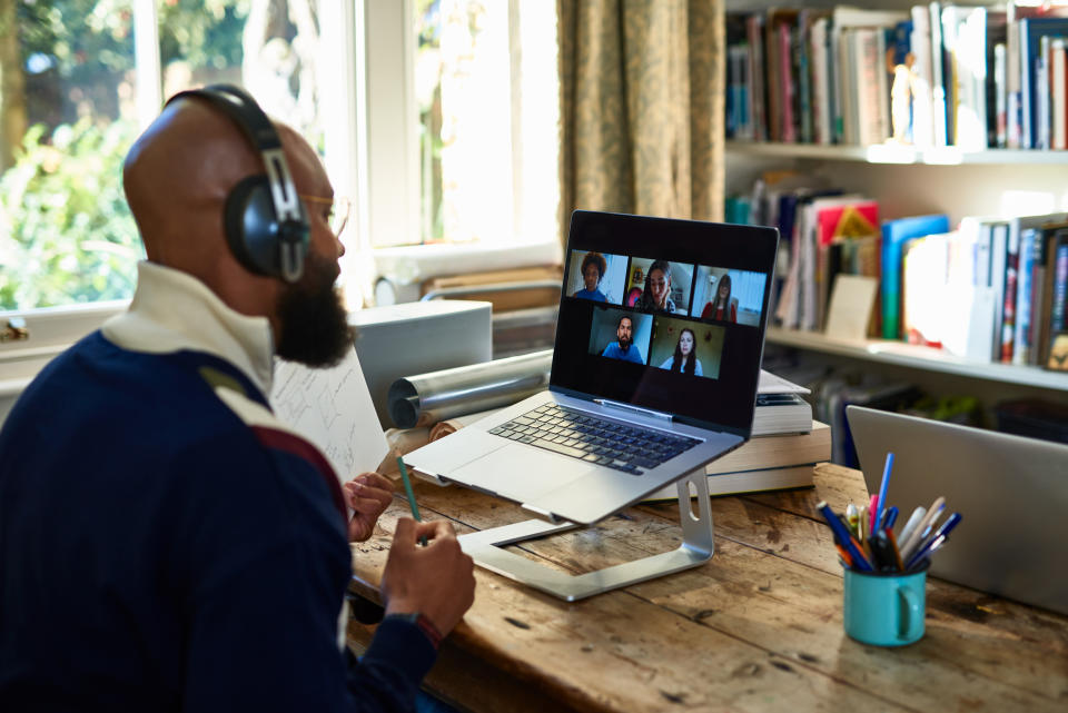 A man wearing headphones participates in a virtual meeting on a laptop at a home office desk cluttered with books and stationery