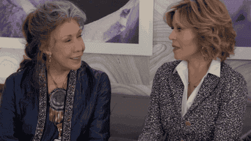 Lily Tomlin and Jane Fonda sit side by side, laughing.