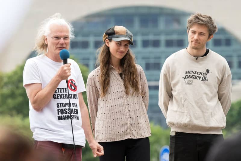 The defendants (L-R) Edmund Schultz, Mirjam Herrmann and Henning Jeschke stand on the stage during a demonstration to mark the indictment by the Neuruppin public prosecutor's office against five members of the Last Generation on charges of "forming a criminal organization". The Federal Chancellery can be seen in the background. Sebastian Christoph Gollnow/dpa