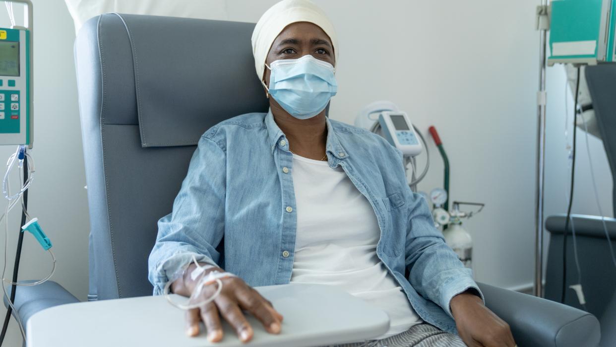  woman with dark brown skin sits in a doctor's office and receives chemotherapy. She is wearing a white head scarf, white undershirt and jean button up, as well as a blue surgical mask 