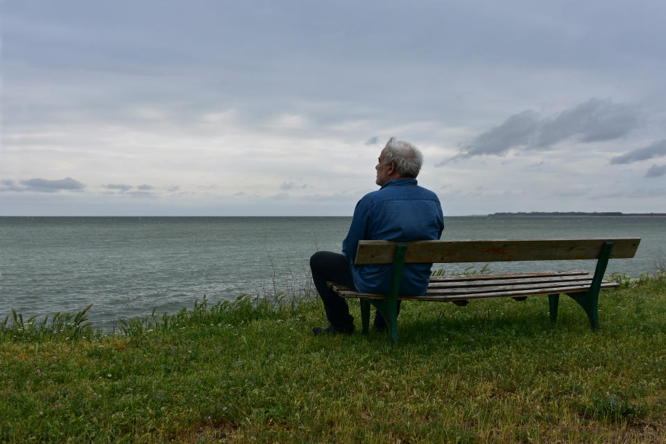 Elderly man sitting on an old wooden bench above the see in a cloudy day, contemplating the nostalgic sea-view