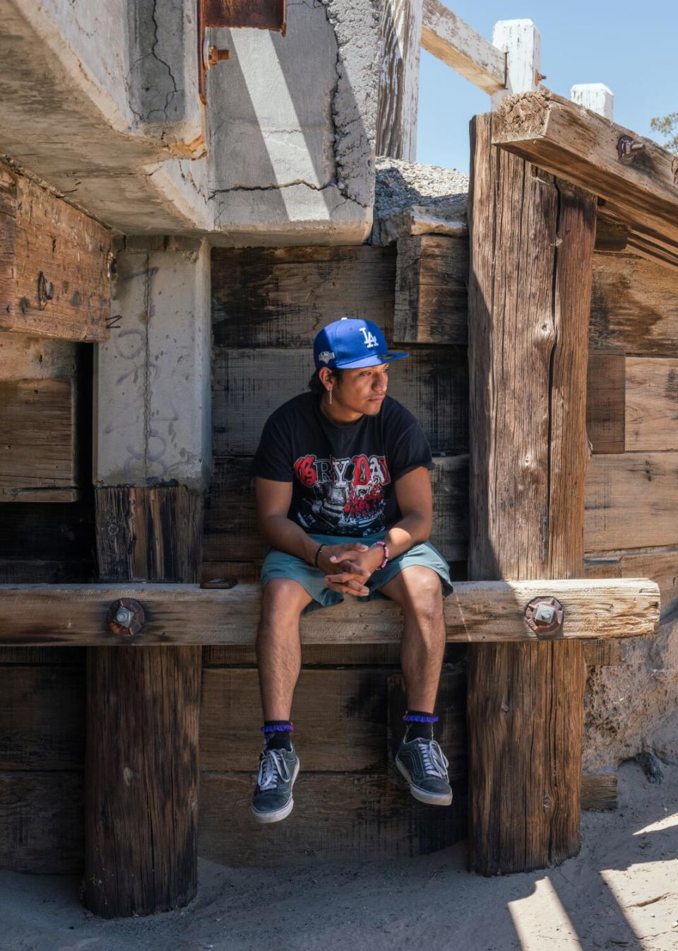 In a vertical full-body frame, a young somber man sits on a wooden beam, in partial shade of an overpass, looking away