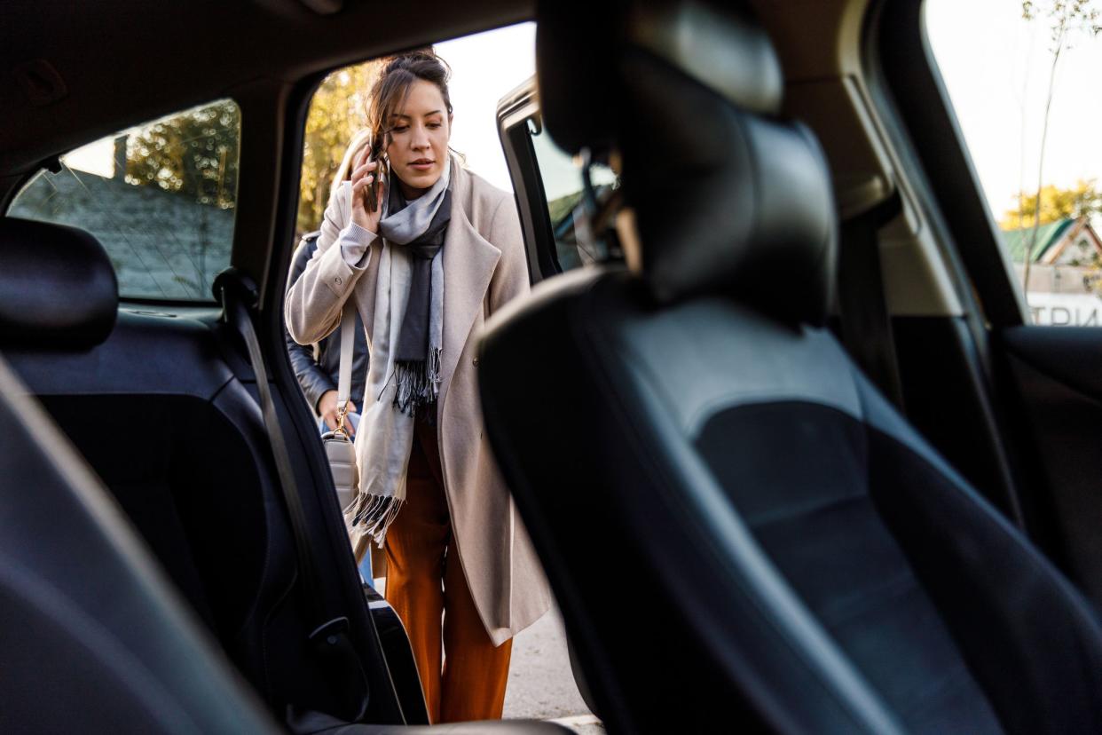 Selective focus shot of smiling young brunette entering a car through the back door and talking on the phone with a friend.
