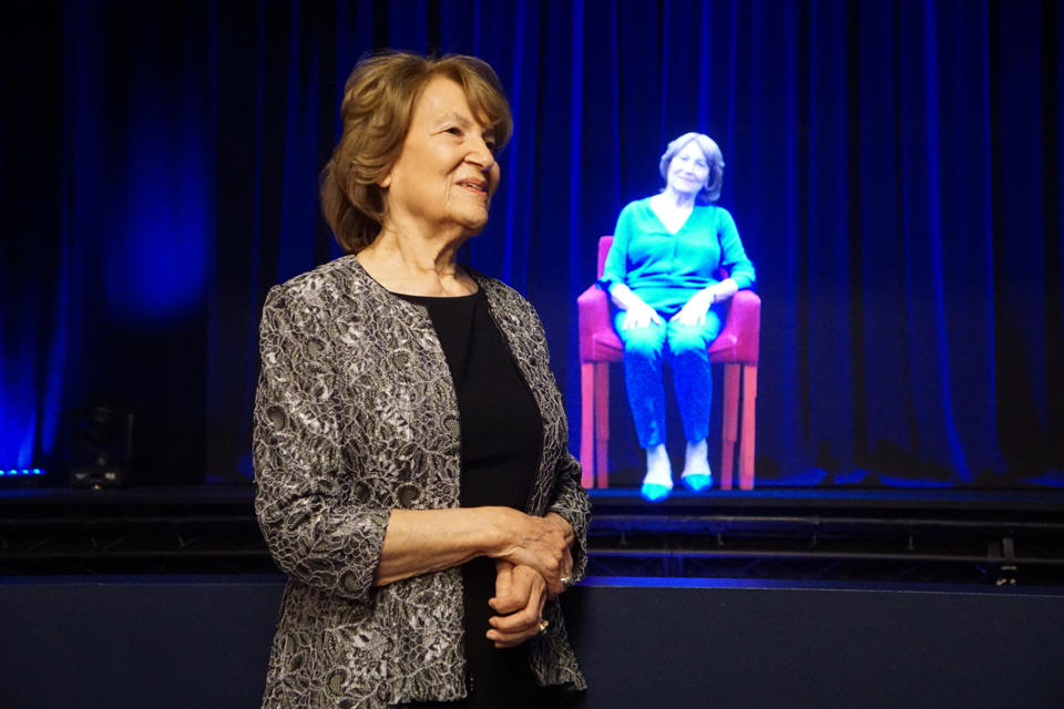 In this October 2017 photo, Holocaust survivor Fritzie Fritzshall stands in front of a hologram of herself at The Abe & Ida Cooper Survivor Stories Experience in the Illinois Holocaust Museum & Education Center in Skokie, Ill. The experience combines high-definition holographic interview recordings and voice recognition technology to enable Holocaust Survivors to tell their deeply moving personal stories and respond to questions from the audience. (Ron Gould/Illinois Holocaust Museum & Education Center via AP)