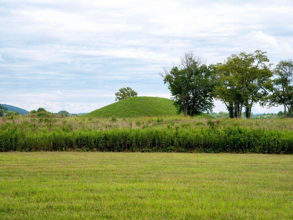 Grass covered prehistoric Hopewell culture burial mound at Siep Earthworks in Ohio under a dramatic cloudy sky with green grass and meadow foreground.