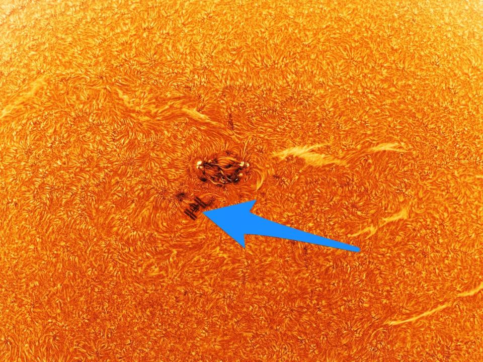 the sun up close turbulent orange surface with a blue arrow pointing the space station tiny silhouette against sunspots