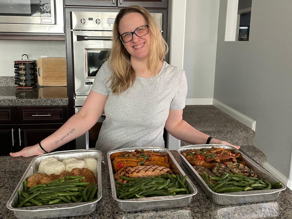 The writer smiles and stands behind a counter with aluminum containers of salmon, chicken, green beans, and other sides in front of her
