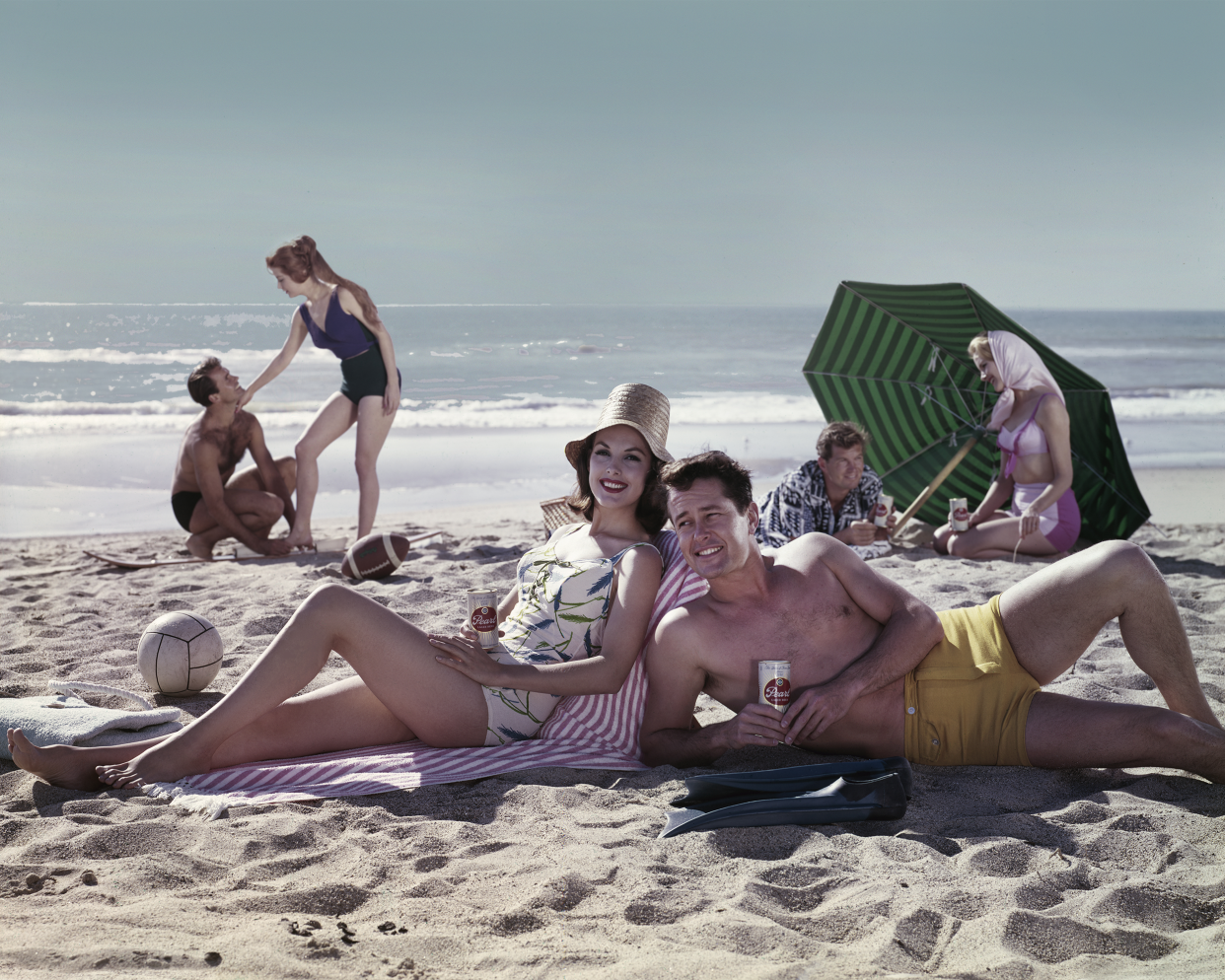 Three couples relax on the beach, with two couples holding Pearl beers in hand, 1960