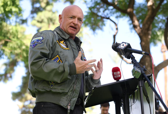 Sen. Mark Kelly, under a tree and wearing a NASA jacket, at the microphone.