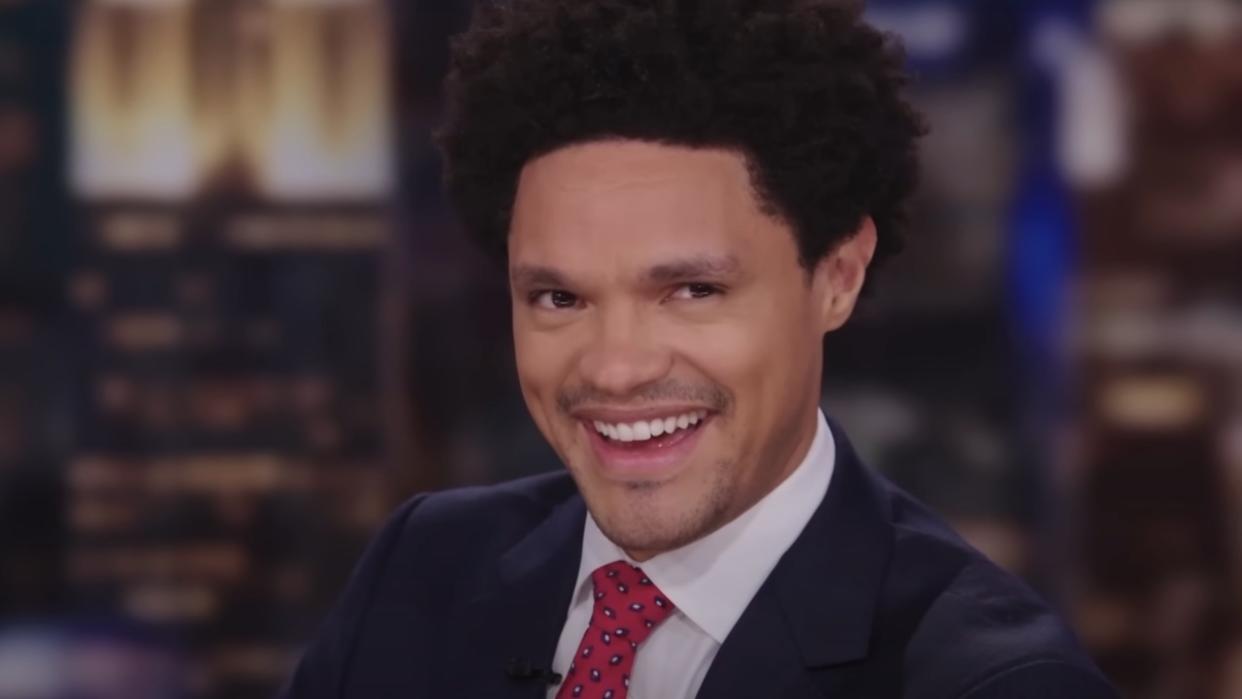  Trevor Noah smiling behind The Daily Show desk 