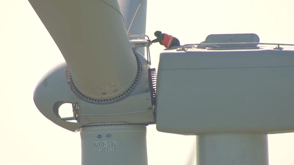 A worker kneels atop a massive wind turbine, and peers into the space between the base and the propeller.