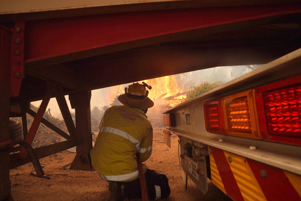 In this photo provided by Department of Fire and Emergency Services, a firefighter shelters under a truck and trailer as he works at a fire near Wooroloo, northeast of Perth, Australia, Tuesday, Feb. 2, 2021. An out-of-control wildfire burning northeast of the Australian west coast city of Perth has destroyed dozens of homes and was threatening more. (Evan Collis/DFES via AP)