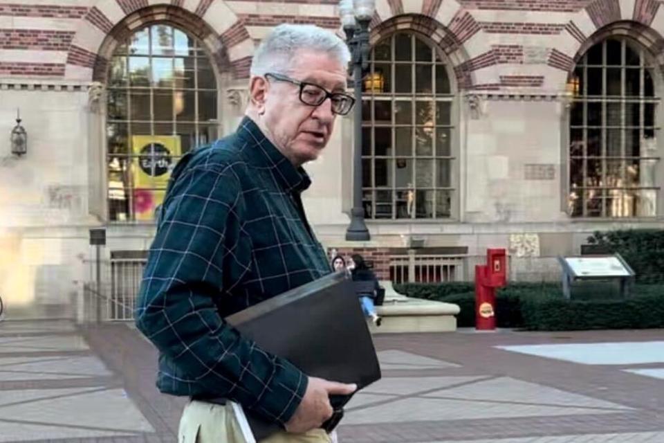 A man with gray hair, in glasses and a plaid green shirt walks near a building