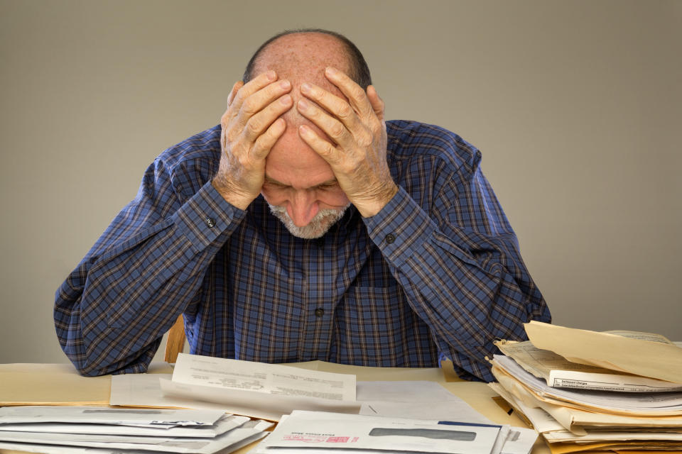 An elderly adult man sitting at a table or desk with stacks of papers and envelopes, hands on his head and looking down. 