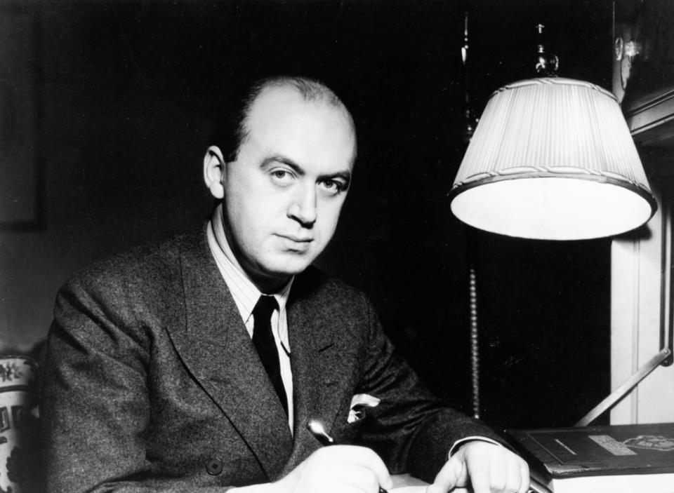 A man in a suit, possibly from a past era, sits at a desk writing with a pen under a lamp