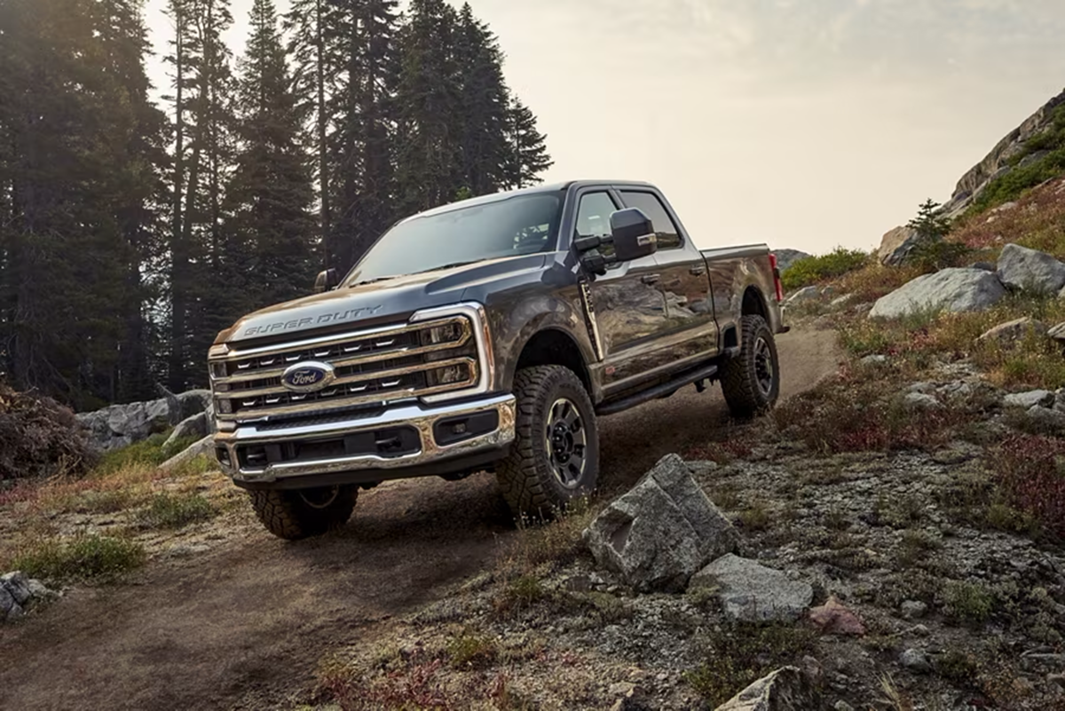 A Ford F-350 Super Duty truck drives down an inclined dirt road surrounded by trees, grass, and boulders.