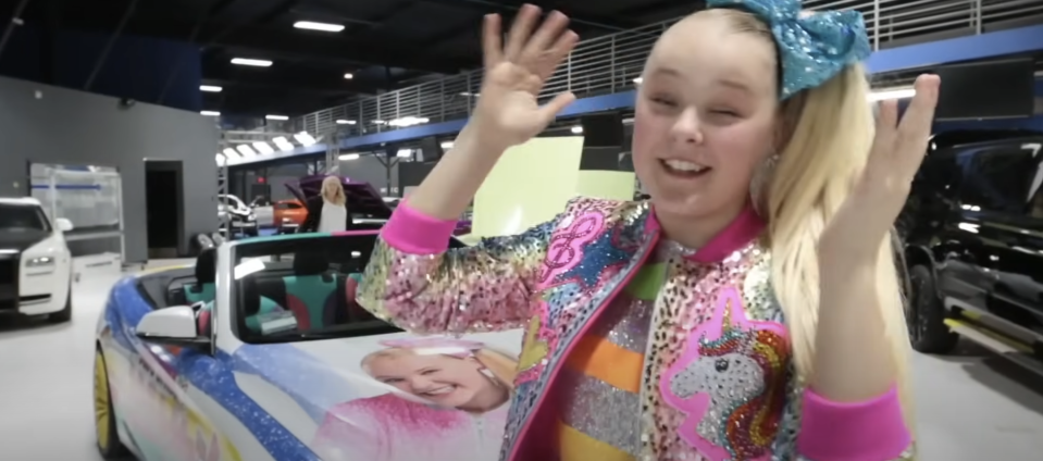 JoJo Siwa in a colorful jacket next to a car with her image on it, waving enthusiastically