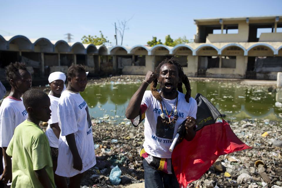 In this Dec. 16, 2018 photo, a man wearing a T-shirt showing former President Jean Bertrand Aristide and clutching a flag associated with the Pitit Dessalines political party decries the government as people gather at a pig farm, once a school, where the victims of a massacre were fed to the animals in the La Saline slum of Port-au-Prince, Haiti. Some residents and local rights groups accuse mid-ranking officials of orchestrating the Nov. 13 attack by a rival gang to intimidate residents and prevent anti-government protests. (AP Photo/Dieu Nalio Chery)