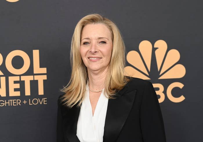 Lisa Kudrow poses in a white blouse and black blazer at the Carol Burnett: 90 Years of Laughter + Love event backdrop