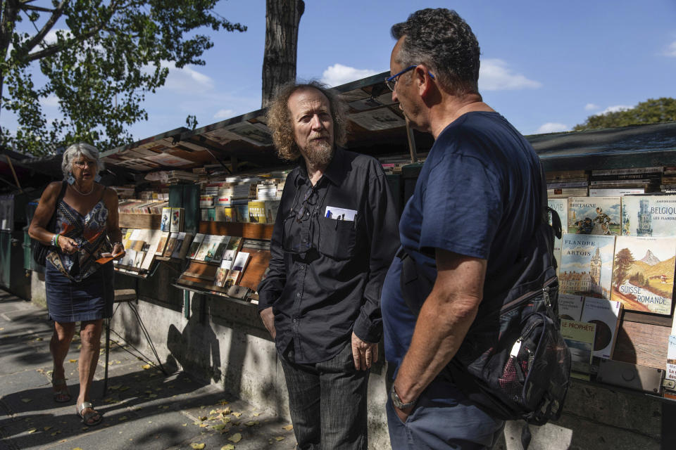 Jerome Callais, left, head of the Paris Booksellers' Cultural Association, talks to a man at his bookseller booth, called "bouquiniste" along the Seine Riverbank in Paris, Tuesday, Aug. 22, 2023. The host city of Paris vowed to deliver an extraordinary grand opening on July 26, 2024, as the ceremony is expected to draw about 600,000 spectators to the Parisian quayside. Citing security measures, the Paris police prefecture ordered on July 25 the removal of 570 stationary boxes out of which booksellers have operated for decades on the quays of the Seine river. (AP Photo/Sophie Garcia)