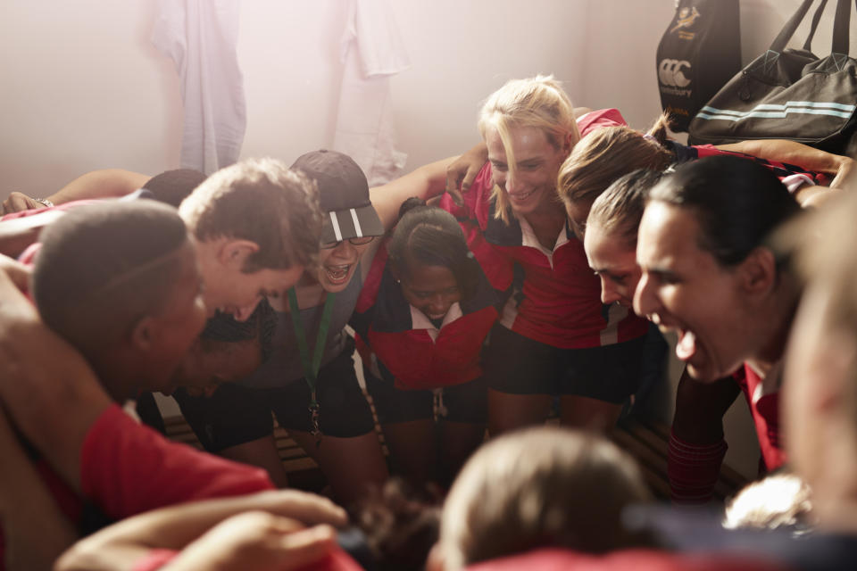 A group of sports team members huddle together enthusiastically in a locker room, smiling and cheering each other on. They are wearing sports uniforms