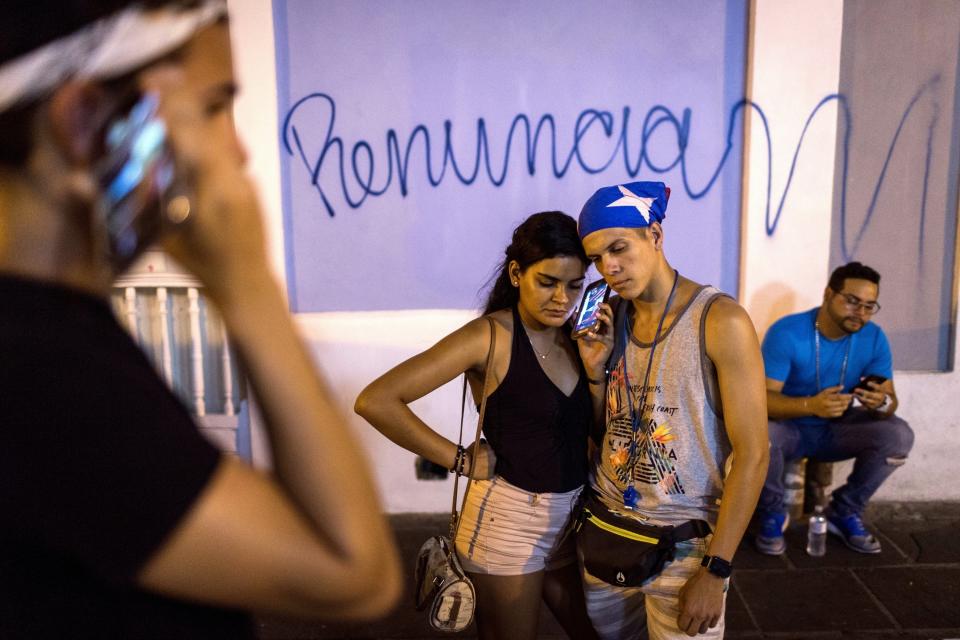 FILE - In this July 24, 2019 file photo, locals listen on a smart phone to the pre-recorded message by Puerto Rico Gov. Ricardo Rossello announcing that he is resigning Aug. 2, after weeks of protests over leaked obscene, misogynistic online chats, in San Juan, Puerto Rico. During weeks of protests preceding the resignation of Gov. Rossello, cellphones across Puerto Rico dinged dozens of times a day as regular citizens and groups including labor unions posted announcements on social media about upcoming protests. (AP Photo/Dennis M. Rivera Pichardo, File)