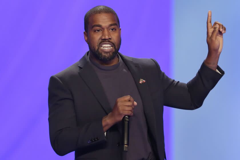 A man onstage holds a microphone in his right hand and raises his left index finger