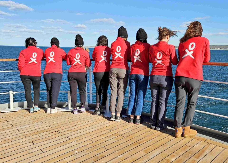 Women in matching red shirts stand on a ship and look at the sea.  Their backs are to the camera.