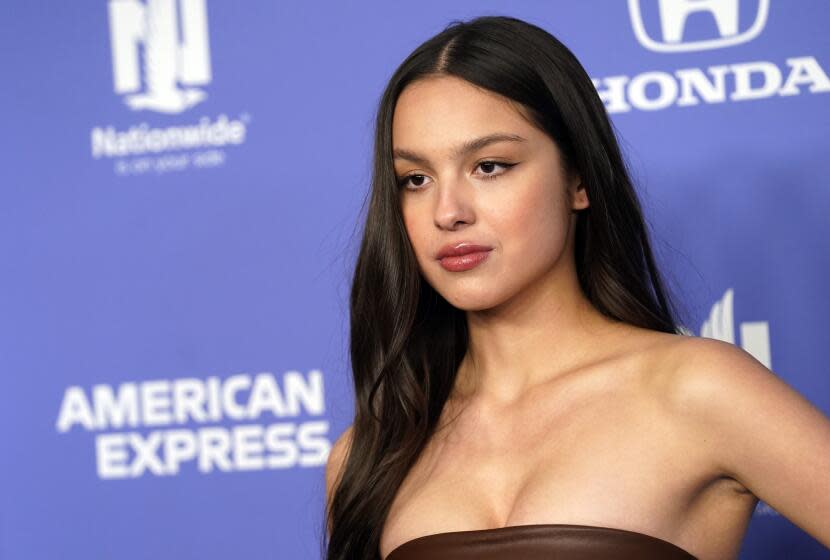 Olivia Rodrigo posing in a sleeveless brown dress against a blue backdrop.