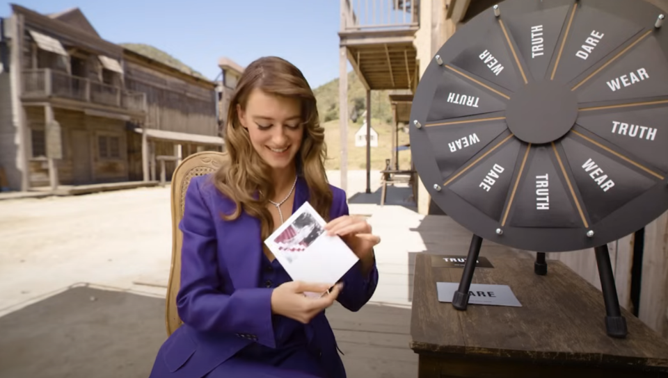 Daisy Edgar-Jones sitting by a spin-the-wheel game, smiling while holding a card, on a western-themed outdoor set. The wheel has options like "Truth" and "Dare."