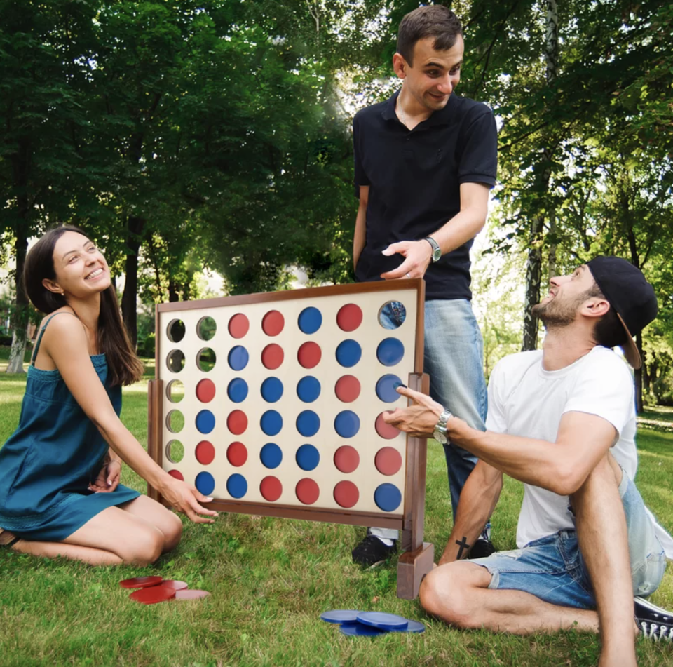 three friends playing red and blue 4-In-A-Row Giant Board Game outside in green park (Photo via Wayfair)