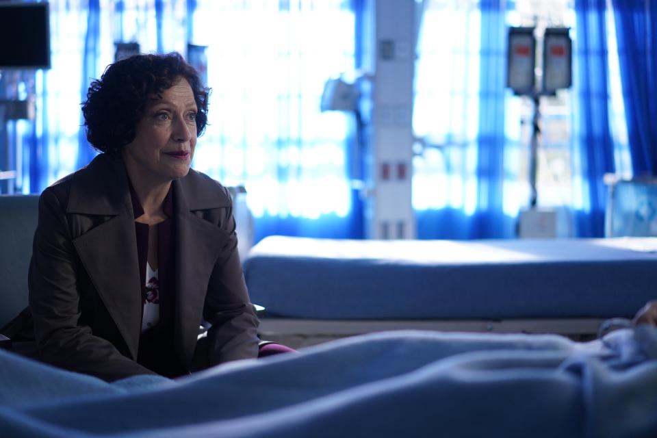 Karin Konoval in blazer sits by a bed in a hospital set, looking concerned in a scene from Big Sky