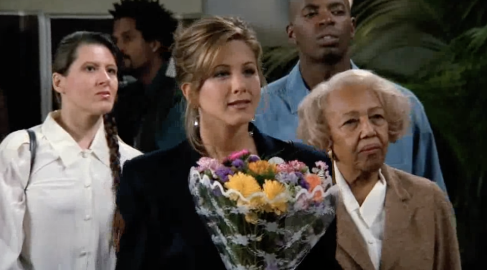 Rachel from Friends holds a bouquet of flowers in an airport, surrounded by people