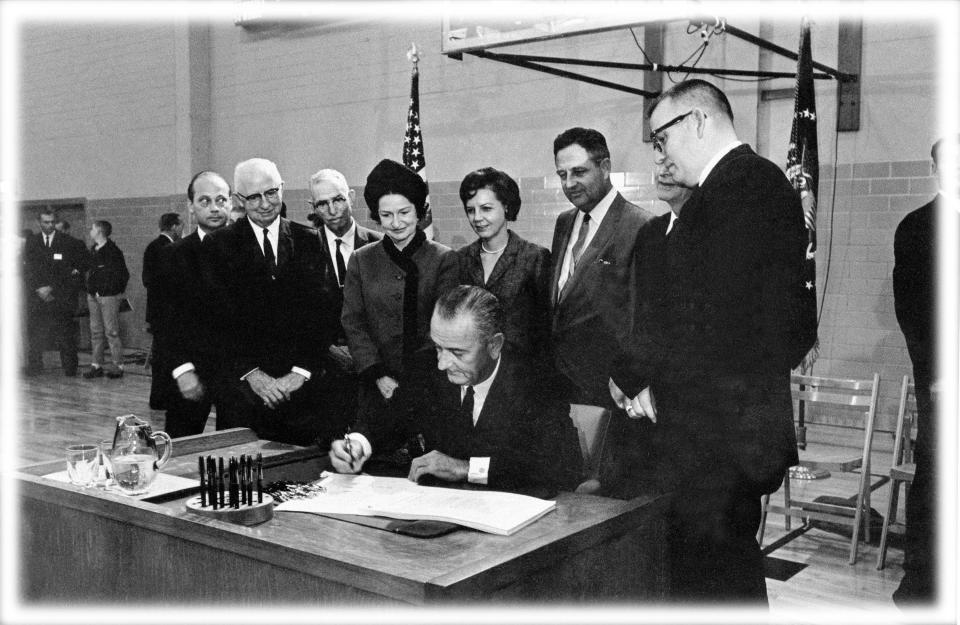 U.S. President Lyndon B. Johnson, seated at the desk he used while a student secretary at Southwest Texas State College 35 years ago, signs the Higher Education Act at San Marcos, Texas, Nov. 8, 1965.  Looking over the president's shoulder is Lady Bird Johnson and to her right are, Mrs. Jake Pickle; Rep. James Jarrell "Jake" Pickle, D-Texas; J.C. Kellam, of Austin, partially hidden; and James H. McCrocklin, president of the college.  The men at extreme left are not identified.  (Photo: AP; digitally enhanced by Yahoo News)