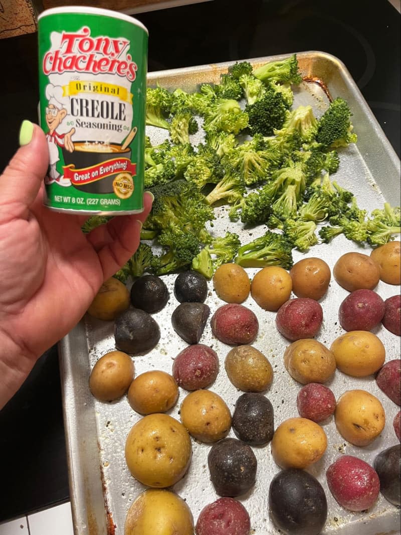 pan of potatoes and broccoli on sheet pan, pre-roasting. Someone holding a container of Tony Chachere's Orginal Creole Seasoning