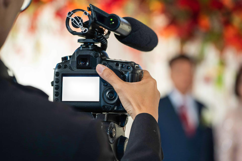 A videographer films a wedding ceremony, focusing on the camera with the bride and groom blurred in the background