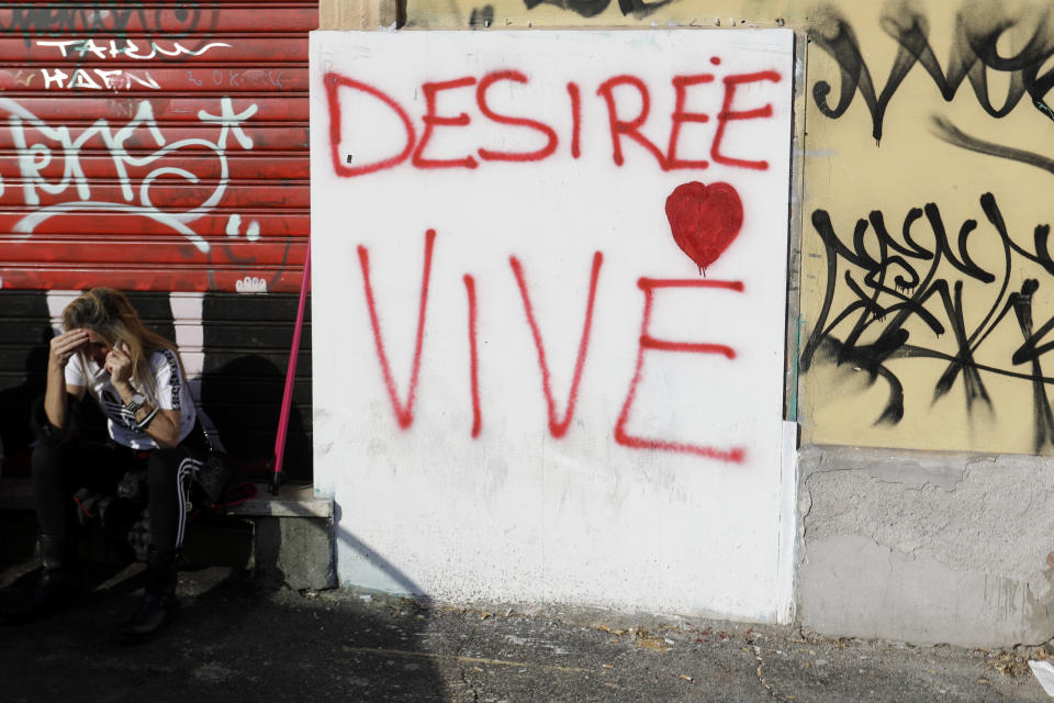 A girl talks on the phone as she sits next to a wall with a writing reading in Italian "Desiree Lives", in Rome, Thursday, Oct. 25, 2018. Italian police have detained three suspects, all immigrants, in the slaying of a teenage girl who was drugged, gang-raped and left in an abandoned building known as a center for drug dealing in Rome. Authorities said Thursday that the three suspects, two Senegalese citizens and one Nigerian, were being held on suspicion of murder, group sexual assault and handing out drugs in the death late last week of 16-year-old Desiree Mariottini. (AP Photo/Gregorio Borgia)