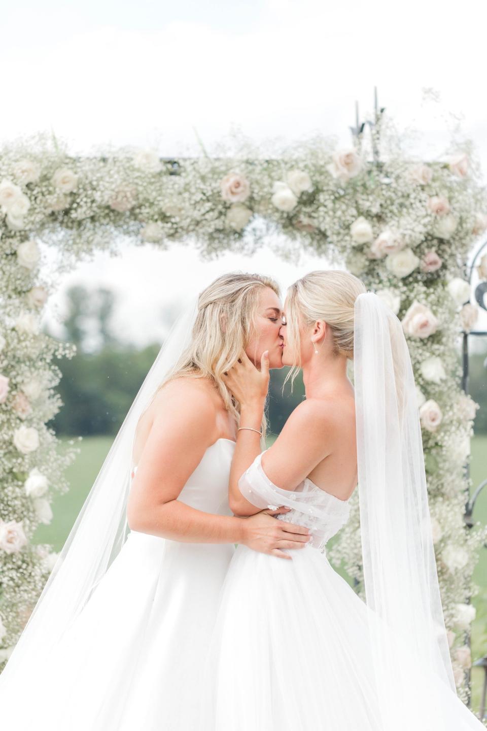 Two brides kiss in front of a white floral archway.