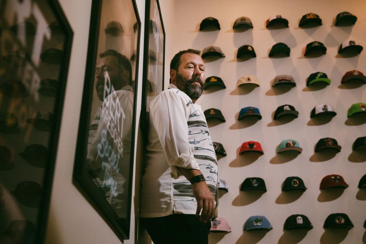 Bert Kreischer stands next to a wall of baseball caps.