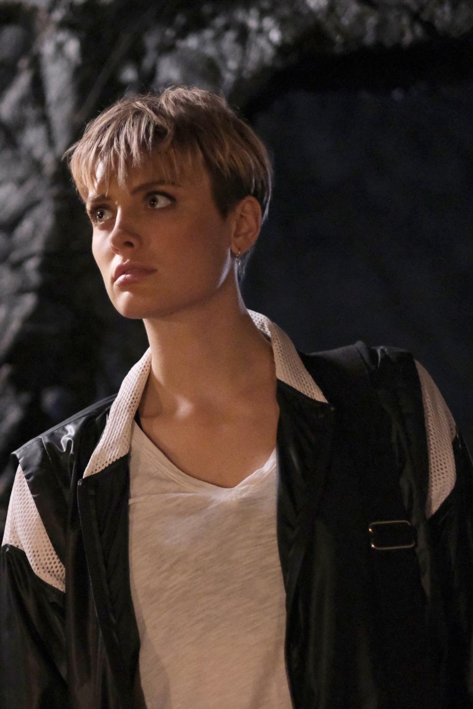 Wallis Daywith short hair in casual attire stands in front of a rocky wall, looking towards the left with a concerned expression
